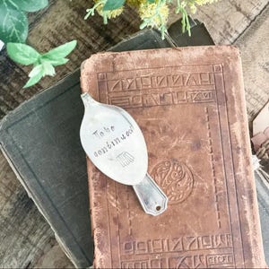 May include: A silver spoon bookmark with the words "To be continued" stamped on the spoon's bowl. The bookmark rests on a vintage, leather-bound book with embossed details. The background includes another book and greenery.