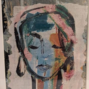 May include: A framed abstract painting featuring a stylized face rendered in thick, expressive brushstrokes. The color palette includes shades of blue, black, pink, and orange, with visible texture on the canvas surface and a white border frame.