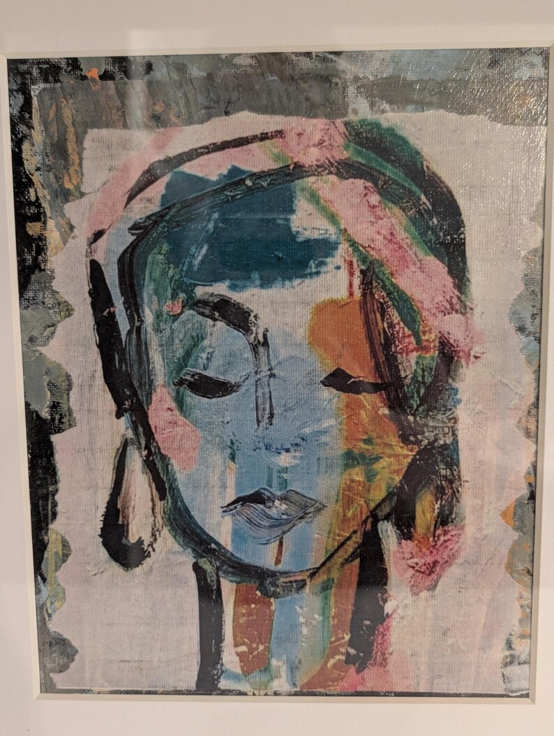 May include: A framed abstract painting featuring a stylized face rendered in thick, expressive brushstrokes. The color palette includes shades of blue, black, pink, and orange, with visible texture on the canvas surface and a white border frame.