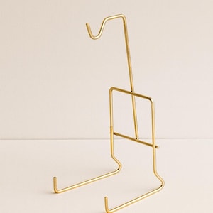 May include: A gold-colored metal display stand with a minimalist design. The stand features a curved top, a rectangular frame, and two horizontal supports at the base. It is designed to hold and display items.