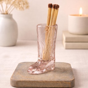 Puede incluir: Un portafósforos transparente de vidrio rosa en forma de bota, lleno de fósforos de madera. El soporte está sobre una pequeña tabla de madera cuadrada. Un jarrón blanco con flores secas y una vela encendida están en el fondo.