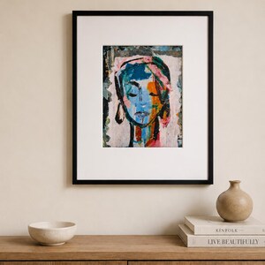 May include: A framed art print featuring an abstract portrait of a face with closed eyes. The artwork uses a vibrant color palette of blue, orange, pink, and black paint strokes on a light background, enclosed in a simple black rectangular frame.