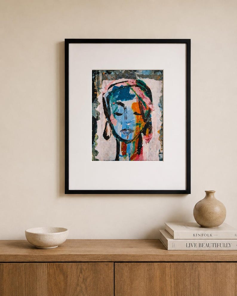 May include: A framed art print featuring an abstract portrait of a face with closed eyes. The artwork uses a vibrant color palette of blue, orange, pink, and black paint strokes on a light background, enclosed in a simple black rectangular frame.