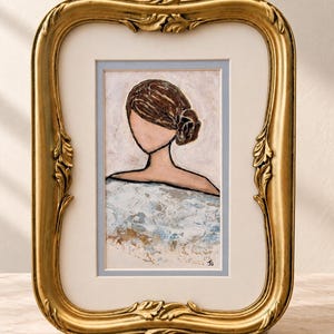 May include: A framed painting of a faceless figure with brown hair in a bun, wearing a textured white and blue garment. The artwork is encased in an ornate, metallic gold frame with leaf-like carvings and a cream-colored mat with a light blue border.