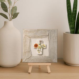 May include: A small, square canvas art piece featuring a white cross adorned with colorful flowers. The cross is centered on a textured, off-white background, framed by a gray, textured border. The artwork is displayed on a miniature wooden easel.