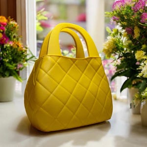 May include: A vibrant yellow handbag featuring a diamond quilted design. It has two curved handles and a structured form. The background shows a window and potted flowers in various colours.