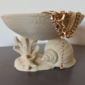 May include: A beige, textured, bowl-shaped trinket dish supported by a base featuring a sculpted coral branch, a starfish, and a seashell. Several gold-toned metal chains, including a link chain and a crystal-accented chain, drape over the edge.