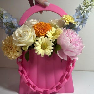 May include: A bright pink handbag-shaped vase filled with a colorful floral arrangement. The flowers include white roses, yellow daisies, orange marigolds, and pink peonies. The vase has a chain strap and a curved handle.