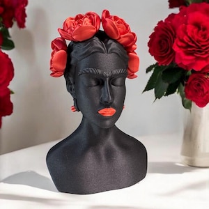 May include: A black bust sculpture of a woman with closed eyes, adorned with a red flower crown and matching earrings. The sculpture has orange lips and is set against a white background with a vase of red roses.