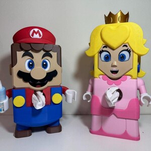 May include: Two character-shaped tissue box holders. One is Mario, with a red hat and blue dungarees. The other is Princess Peach, in a pink dress and a gold crown. Both have tissue openings.