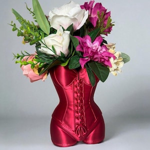 May include: A vibrant red corset-shaped vase, holding a bouquet of mixed flowers and foliage. The vase has a satin sheen and a detailed lace-up front. The floral arrangement includes white roses, pink peonies, and magenta dahlias.