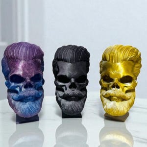 May include: Three decorative skull sculptures with sculpted hair and beards. The skulls are in purple, black, and gold. Each skull has a unique colour and finish, adding a gothic flair to any space.