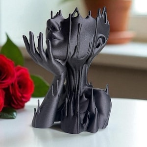May include: A black sculpture of a human face and hands, with a unique, dripped design. The sculpture is resting on a white surface, with red roses in the background. The sculpture's design is a modern take on a bust.