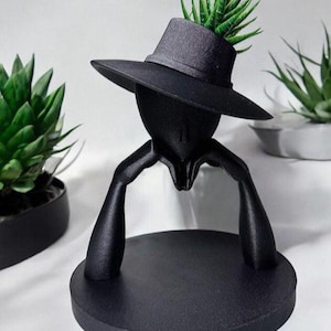 May include: A black decorative planter shaped like a person wearing a hat, with arms resting on a circular base. The hat holds a green succulent plant. The planter is made of a matte black material.
