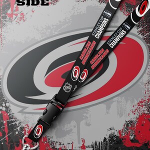 May include: A black lanyard featuring the Carolina Hurricanes logo, the text "Stanley Cup Champions," and the NHL logo. The strap includes a black plastic side-release buckle and a silver-toned metal swivel lobster clasp at the bottom end.
