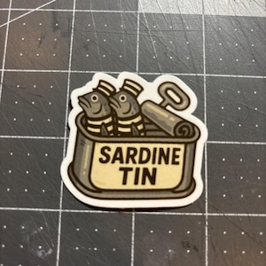 May include: A sticker depicting a sardine tin with two cartoon fish wearing hats. The tin is labelled "SARDINE TIN" in bold letters, and a can opener is shown beside it. The sticker has a white border.
