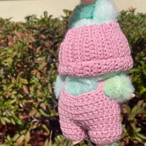 May include: A handmade doll wearing a pink crocheted hat and overalls. The doll has a mint green fluffy head and body, with matching fluffy accents on the arms and legs. The doll is held up by a green loop.