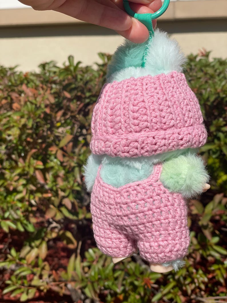 May include: A handmade doll wearing a pink crocheted hat and overalls. The doll has a mint green fluffy head and body, with matching fluffy accents on the arms and legs. The doll is held up by a green loop.