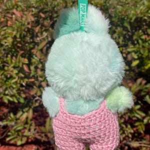 May include: A plush, light blue and green bunny keychain with a pink crocheted overall. The keychain has a green ring and a green tag that reads "POP MART". The bunny's fur is soft and fluffy.