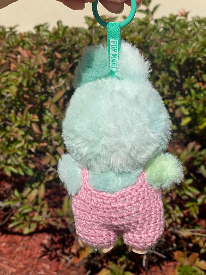 May include: A plush, light blue and green bunny keychain with a pink crocheted overall. The keychain has a green ring and a green tag that reads "POP MART". The bunny's fur is soft and fluffy.