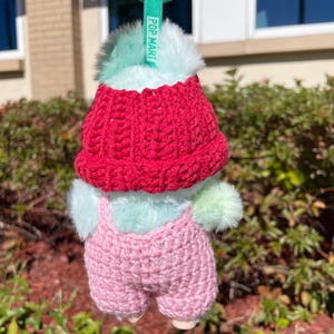 May include: A small, plush toy with a red knitted hat and pink overalls. The toy has a mint green body and fluffy accents. A green tag with the text "POP MART" is attached to a green keyring.