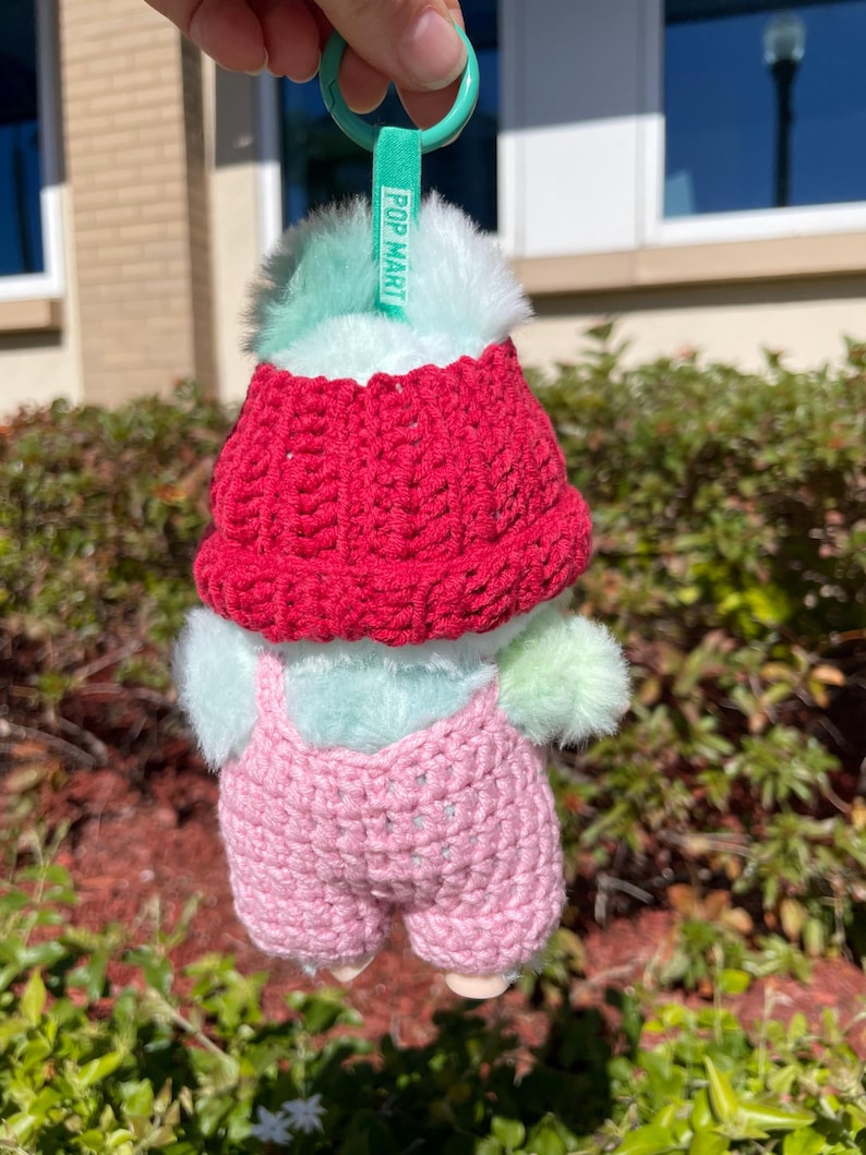 May include: A small, plush toy with a red knitted hat and pink overalls. The toy has a mint green body and fluffy accents. A green tag with the text "POP MART" is attached to a green keyring.