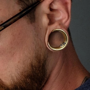 Puede incluir: Un medidor de oreja de color dorado, también conocido como tapón o túnel, se usa en un lóbulo de la oreja estirado. El medidor tiene un diseño de media luna con pequeños detalles grabados. La persona de la imagen tiene barba y usa gafas.