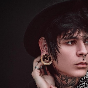May include: Gold-coloured ear gauges with a snake design. The gauges are circular and feature a detailed snake coiled within the hoop. The person in the image is wearing a black hat and has multiple facial piercings.