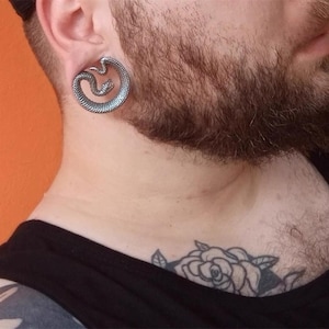 Può includere: Un dilatatore per orecchio color argento con un design dettagliato a forma di serpente arrotolato. Il serpente è avvolto in una forma circolare, creando un accessorio unico e accattivante. Il dilatatore è indossato nell'orecchio.