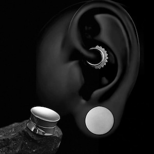May include: A black and white image of body jewelry. A silver-colored hinged hoop earring is in the ear's cartilage. A silver-colored plug and a flat, round plug are also visible. The background is black.