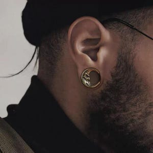 May include: A gold-colored ear plug with a crescent moon design. The moon features a detailed face carved into the surface. The plug is circular and fits into a stretched earlobe, showcasing a unique body modification.