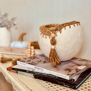 Peut inclure: Un bol décoratif blanc texturé avec un bord crocheté marron et une gland pendante. Le bol est posé sur une pile de livres avec le mot "Félicité" visible. La scène évoque une esthétique chaleureuse et rustique.