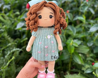May include: A handmade crochet doll with brown curly hair and a light blue bow. The doll wears a light blue dress with pink heart accents and pink shoes. The doll's name, "Erie", is embroidered on the dress.
