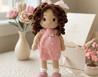 May include: A handmade crochet doll with curly brown hair, a light pink ribbon, and a light pink dress featuring small floral embroidery. The doll has tan skin, white socks, and pink shoes, standing upright with a soft, textured yarn construction.
