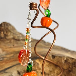 Peut inclure: Une paire de boucles d'oreilles sur le th&egrave;me de l'automne. Chaque boucle d'oreille pr&eacute;sente un petit charme de citrouille orange avec une tige verte, une feuille orange translucide et des perles color&eacute;es. Les boucles d'oreilles sont suspendues &agrave; un crochet et une cha&icirc;ne en argent.