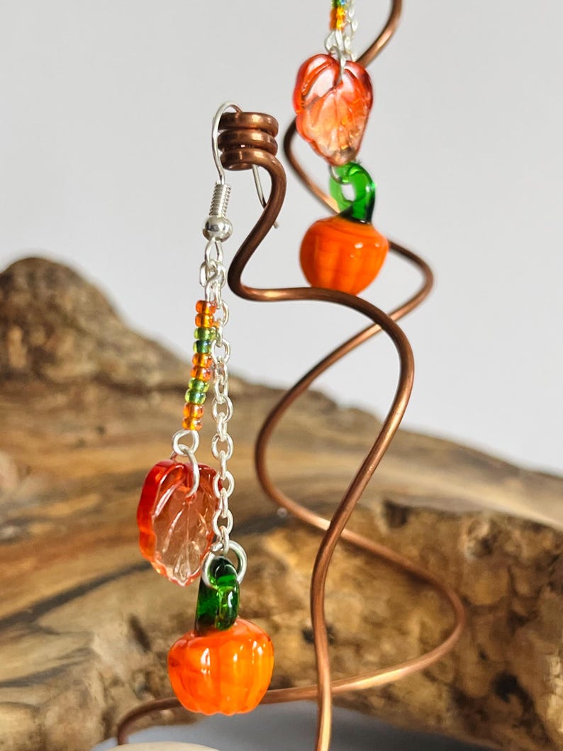 Peut inclure: Une paire de boucles d'oreilles sur le th&egrave;me de l'automne. Chaque boucle d'oreille pr&eacute;sente un petit charme de citrouille orange avec une tige verte, une feuille orange translucide et des perles color&eacute;es. Les boucles d'oreilles sont suspendues &agrave; un crochet et une cha&icirc;ne en argent.