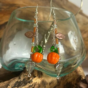 Peut inclure: Boucles d'oreilles faites &agrave; la main avec des perles de citrouille orange, des anneaux en verre vert et des breloques en forme de feuille. Les boucles d'oreilles ont des crochets et des cha&icirc;nes de couleur argent&eacute;e, avec des accents de perles orange et vertes.