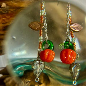 Peut inclure: Boucles d'oreilles faites &agrave; la main avec des breloques citrouille orange, des accents verts et des pendentifs en forme de feuille argent&eacute;e. Les boucles d'oreilles ont un design perl&eacute; avec des perles cuivr&eacute;es et vertes, suspendues &agrave; des cha&icirc;nes argent&eacute;es.