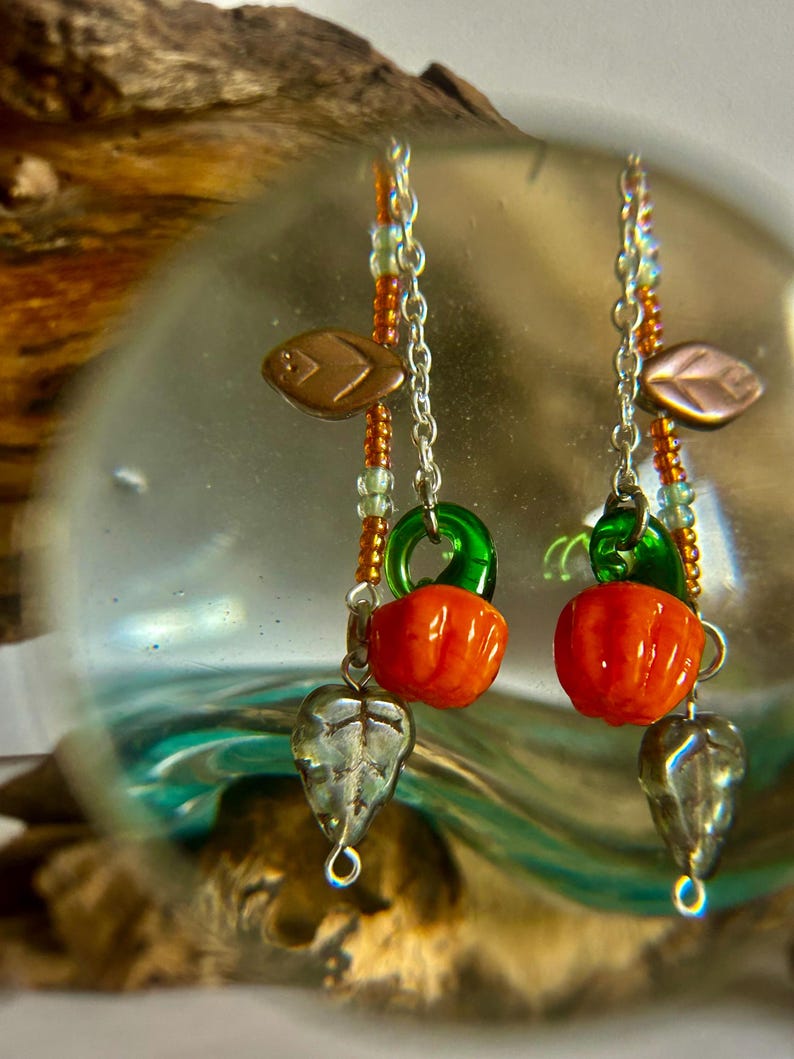 Peut inclure: Boucles d'oreilles faites &agrave; la main avec des breloques citrouille orange, des accents verts et des pendentifs en forme de feuille argent&eacute;e. Les boucles d'oreilles ont un design perl&eacute; avec des perles cuivr&eacute;es et vertes, suspendues &agrave; des cha&icirc;nes argent&eacute;es.
