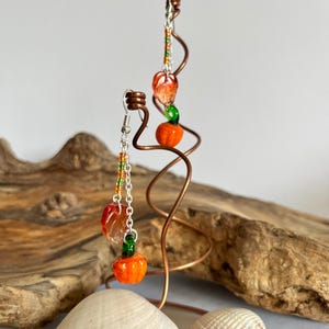 Peut inclure: Boucles d'oreilles faites &agrave; la main avec des breloques en forme de citrouille et de feuille orange. Les boucles d'oreilles sont suspendues &agrave; des crochets et des cha&icirc;nes de couleur argent&eacute;e, avec des perles color&eacute;es. Les breloques sont expos&eacute;es sur une sculpture en fil de cuivre, avec des coquillages au premier plan.