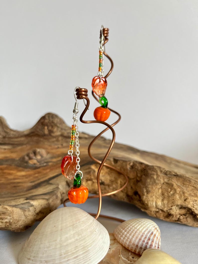 Peut inclure: Boucles d'oreilles faites &agrave; la main avec des breloques en forme de citrouille et de feuille orange. Les boucles d'oreilles sont suspendues &agrave; des crochets et des cha&icirc;nes de couleur argent&eacute;e, avec des perles color&eacute;es. Les breloques sont expos&eacute;es sur une sculpture en fil de cuivre, avec des coquillages au premier plan.