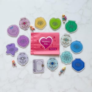 May include: A selection of vintage Polly Pocket compacts in diverse shapes and colours, such as hearts, stars, and shells, alongside miniature figures and a pink mystery bag with the text "mystery bag" and "Open it up".