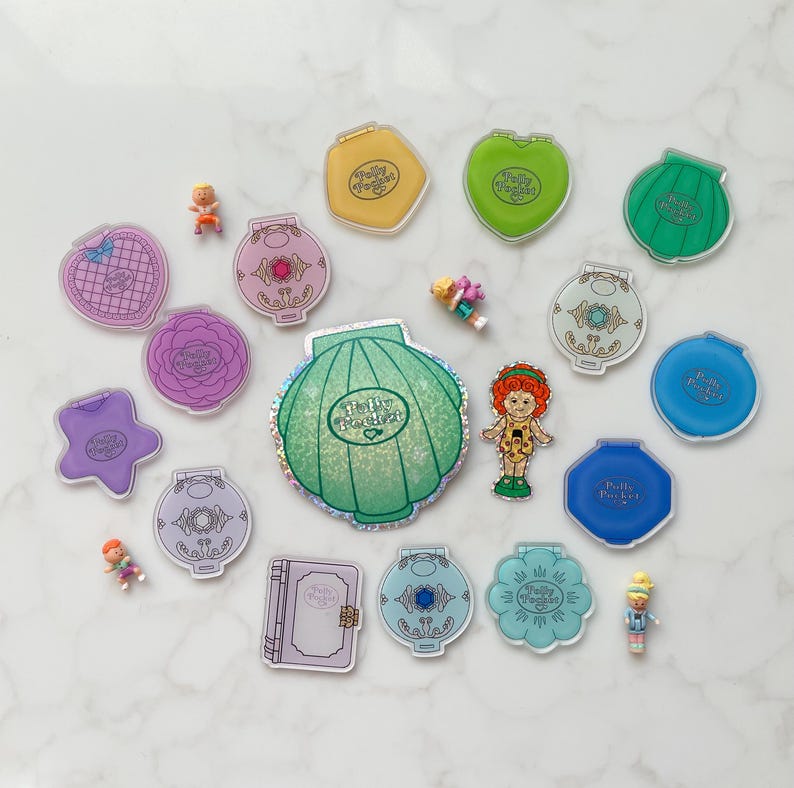 May include: A selection of vintage Polly Pocket toys, including various compacts in shapes such as hearts, shells, and stars, alongside miniature figures. The compacts are in shades of pink, blue, green, and yellow, with detailed designs and the Polly Pocket logo.