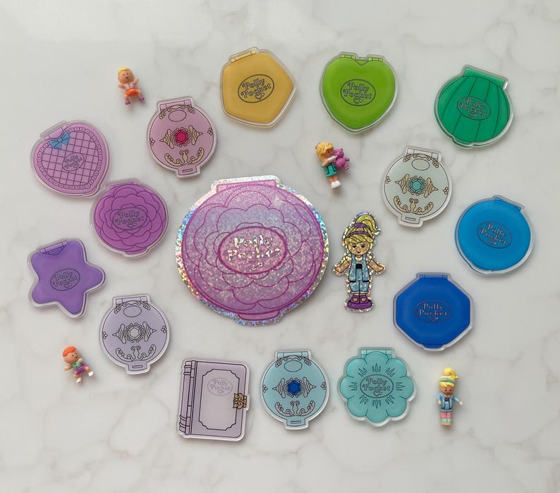 May include: A selection of vintage Polly Pocket toys in diverse shapes and colours, including compacts shaped like flowers, hearts, and shells. Several miniature figures are scattered among the compacts. The Polly Pocket logo is visible on many of the toys.