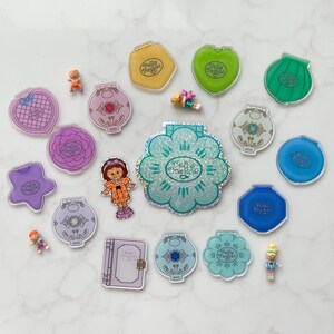 May include: A selection of vintage Polly Pocket toys in diverse shapes and colours, including compacts shaped like flowers, hearts, and stars. Several miniature figures are scattered around the toys, along with a paper doll. The toys are arranged on a white marble surface.