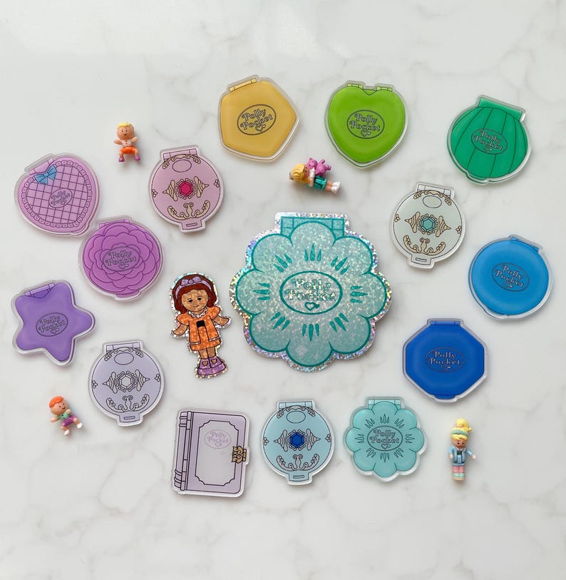 May include: A selection of vintage Polly Pocket toys in diverse shapes and colours, including compacts shaped like flowers, hearts, and stars. Several miniature figures are scattered around the toys, along with a paper doll. The toys are arranged on a white marble surface.