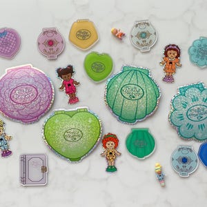 May include: A selection of vintage Polly Pocket toys, including compacts in flower, heart, and shell shapes. The toys are in pink, green, yellow, and blue, with small figurines. Measurements are in centimetres.