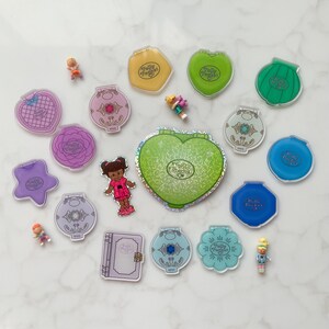 May include: A selection of vintage Polly Pocket toys in diverse shapes and colours, including hearts, stars, and flowers. Several miniature dolls are scattered among the compacts. The Polly Pocket logo is visible on many of the items.