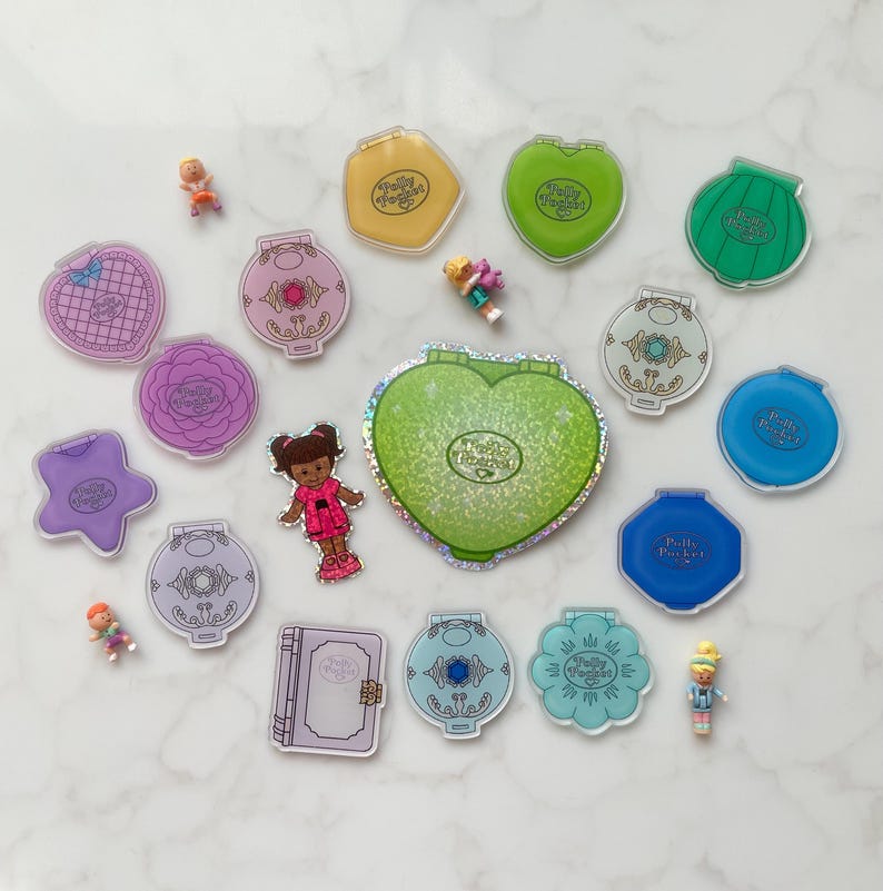 May include: A selection of vintage Polly Pocket toys in diverse shapes and colours, including hearts, stars, and flowers. Several miniature dolls are scattered among the compacts. The Polly Pocket logo is visible on many of the items.