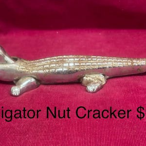 May include: A silver-colored alligator nutcracker. The nutcracker is in the shape of an alligator, with its mouth open. The alligator is lying on a red surface.